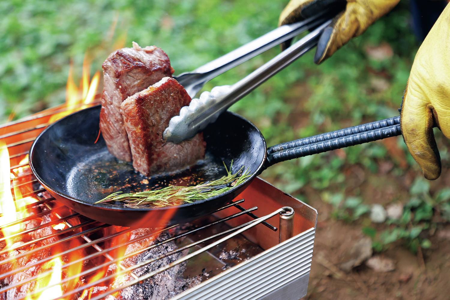 焚火台の上に置いたフライパンで厚切りの肉を焼いている