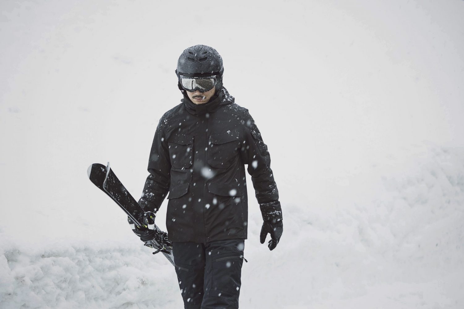 「F70 URBAN EXPLORER JACKET」を着用して雪の中をスキー板を持って歩く