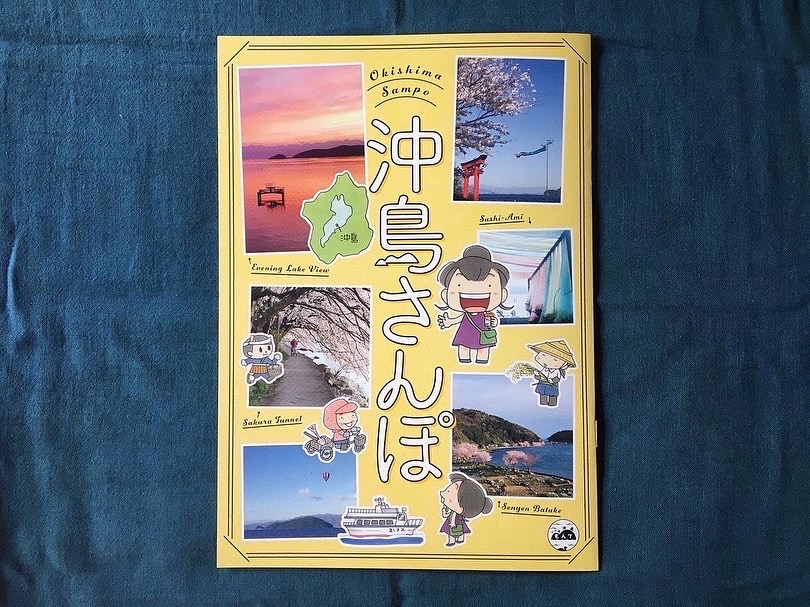 うっすら緑がかった布地の上に黄色地に白字で「沖島さんぽ」と書かれた冊子。文字の周囲には、沖島の四季折々の写真が6枚並べられている