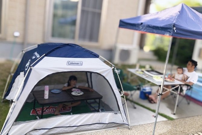 自宅前の駐車場にテント、タープテント、机、プールを立ててBBQをする親子。