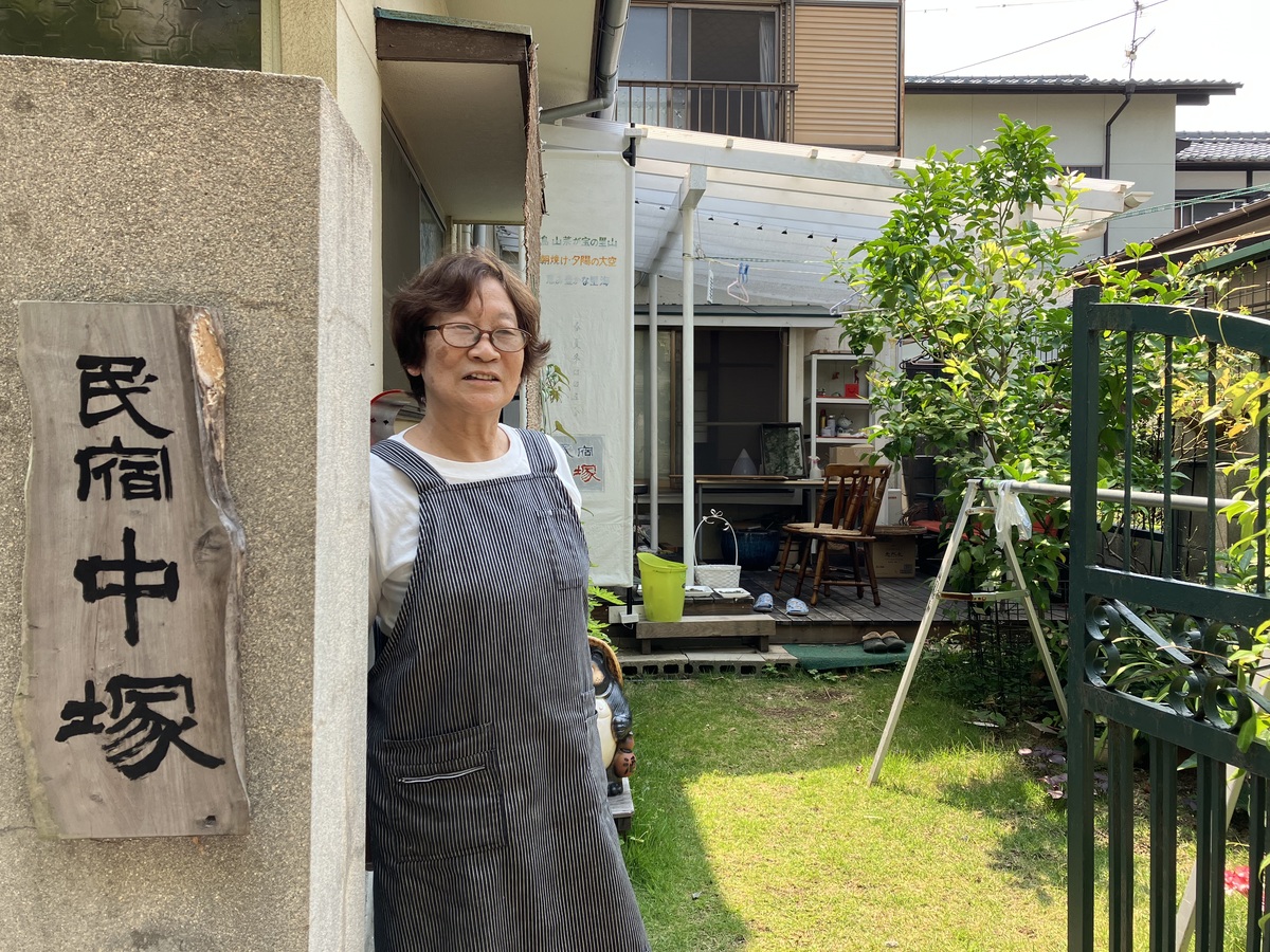 「民宿中塚」と書かれた看板がある宿の入口に微笑んで佇む女将のいつこさん