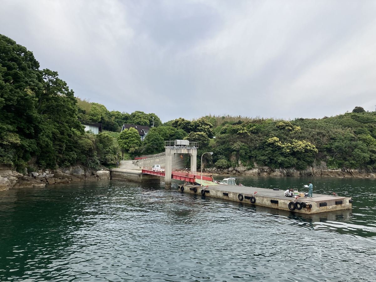 島の港。桟橋の向こうには小さな坂。その周りは木々が生い茂る。木々の合間に建物が少し見える。