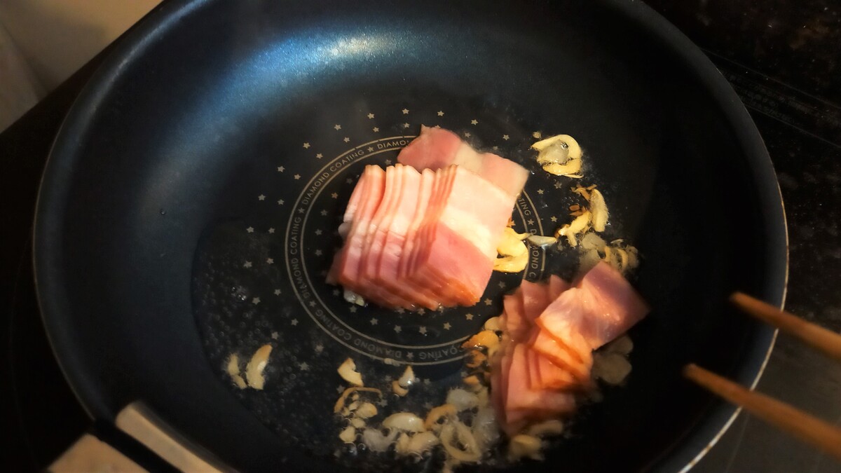 にんにくを炒めているフライパンの中にベーコンを入れている。