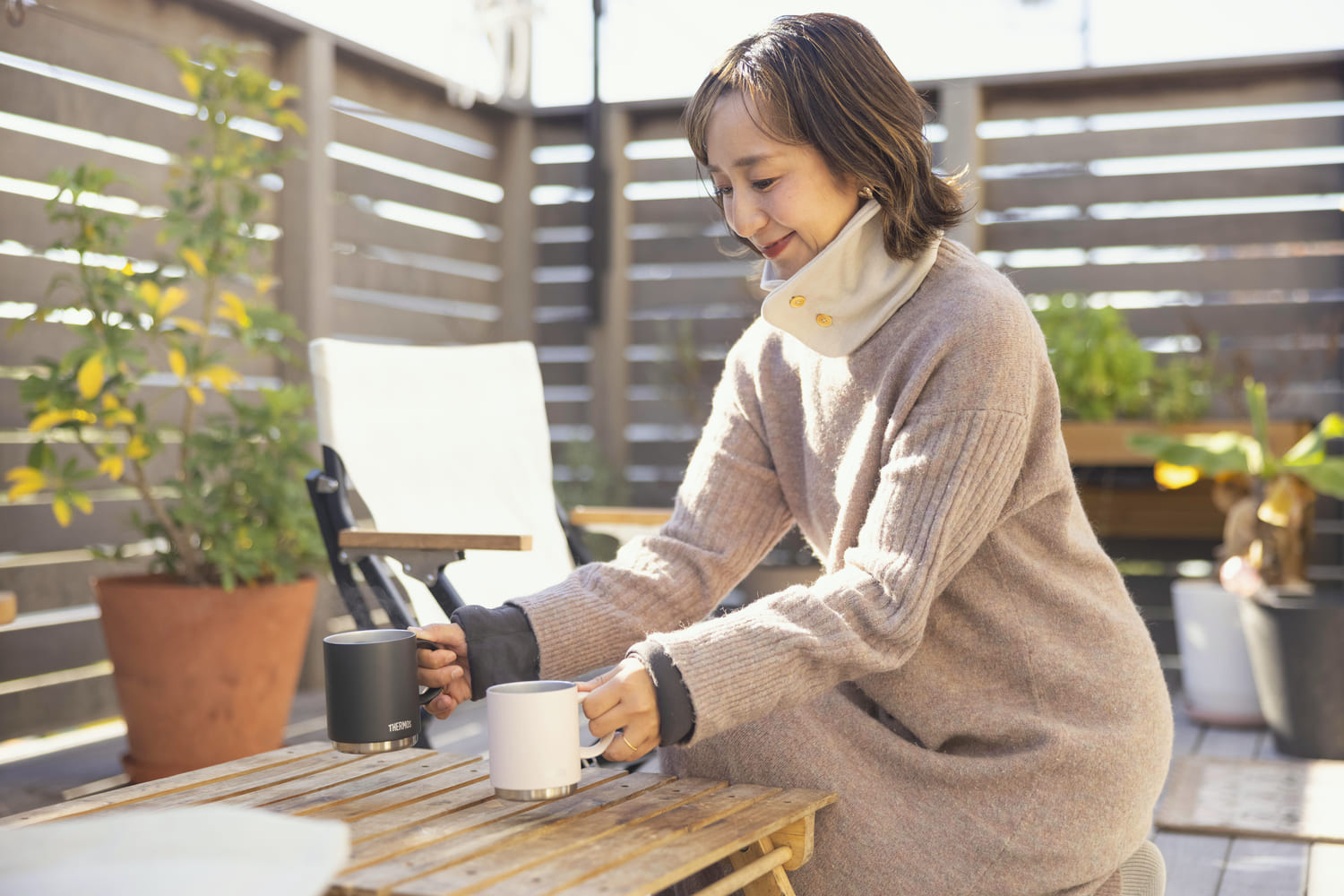 ネックウォーマーを着用し、コーヒーをベランダのテーブルに並べる女性