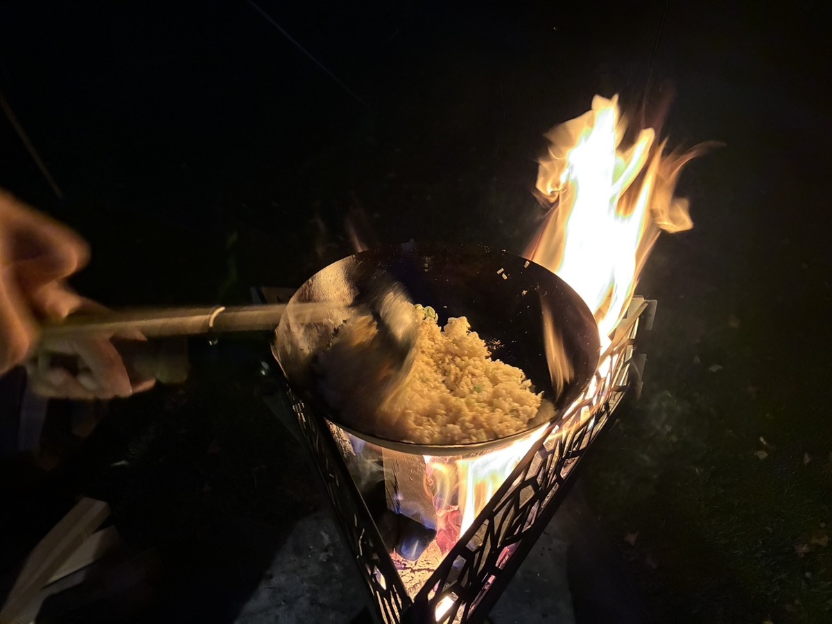 焚き火の炎に包まれながら中華鍋でチャーハンを炒めている様子