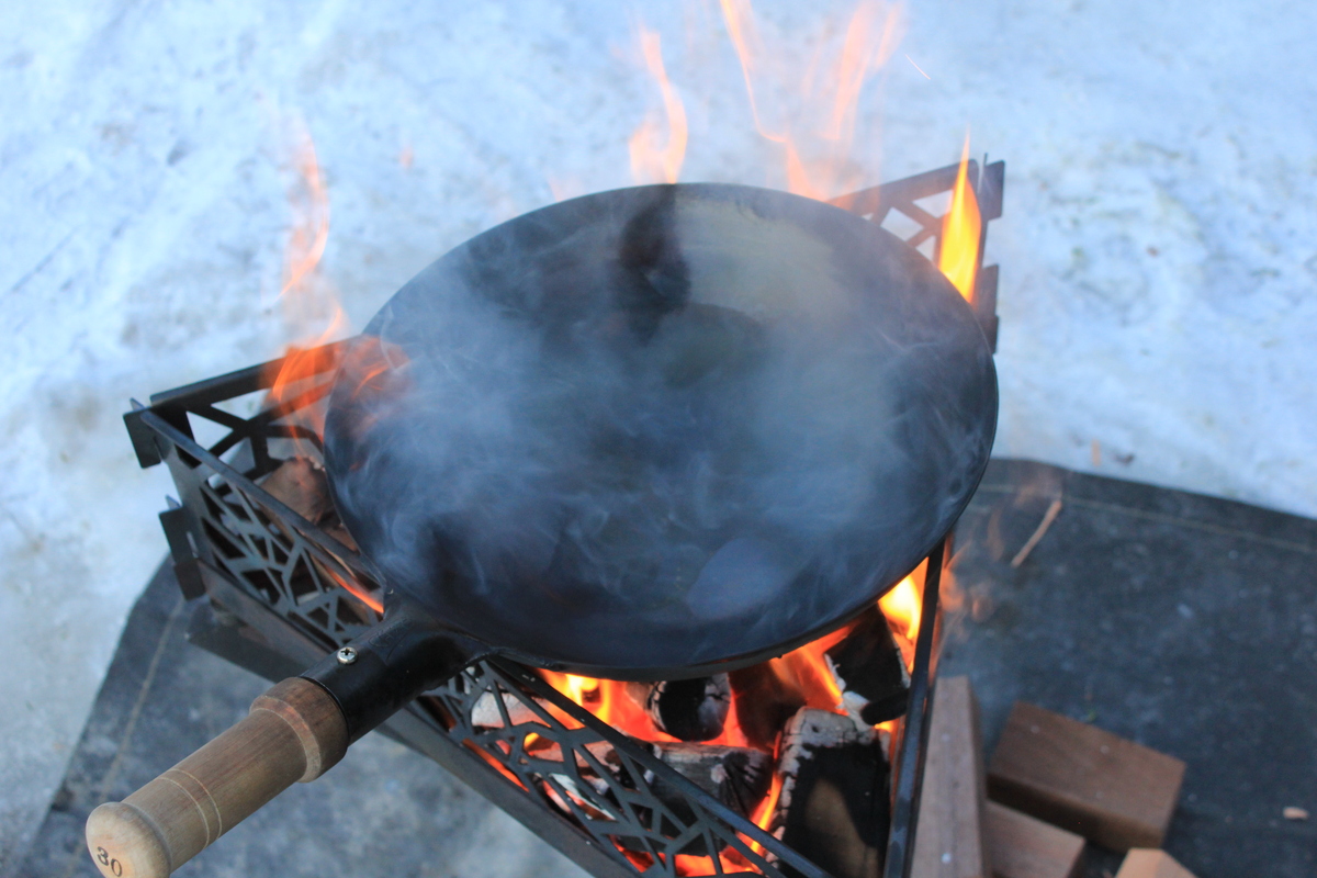 焚き火の上で空焼きされ、白い煙が立ち上る鉄製中華鍋