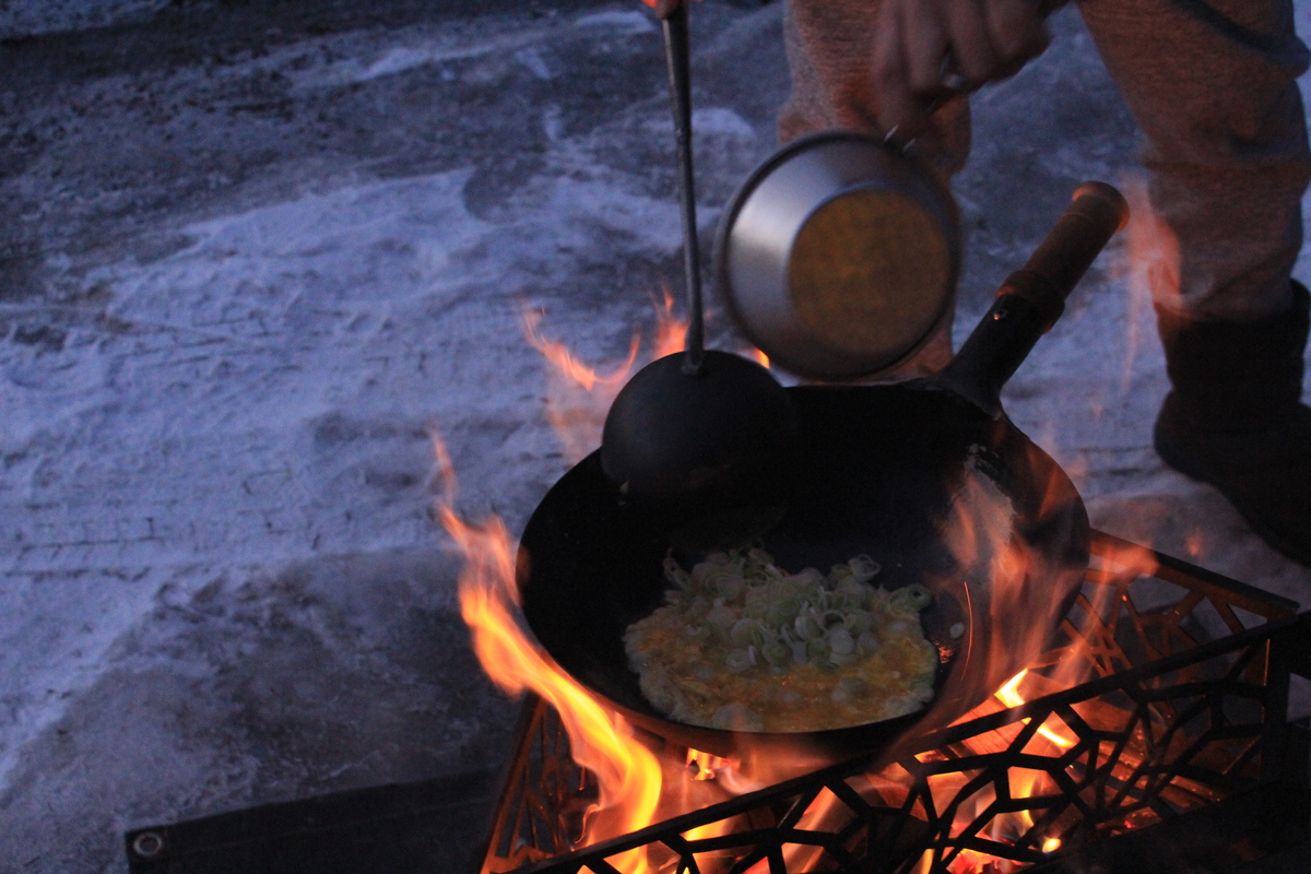 焚き火にかけた中華鍋に卵と具材を加えて調理している様子