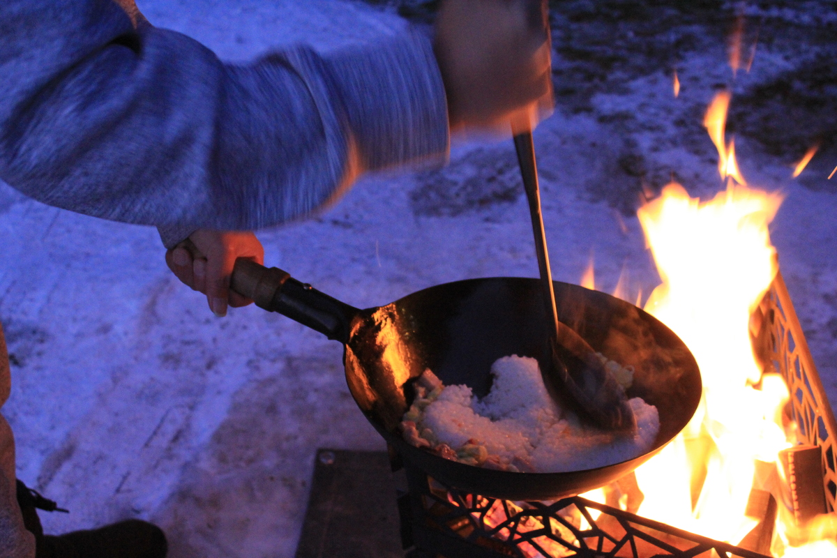 焚き火の強火で中華鍋に入れたごはんをおたまでほぐしている調理シーン